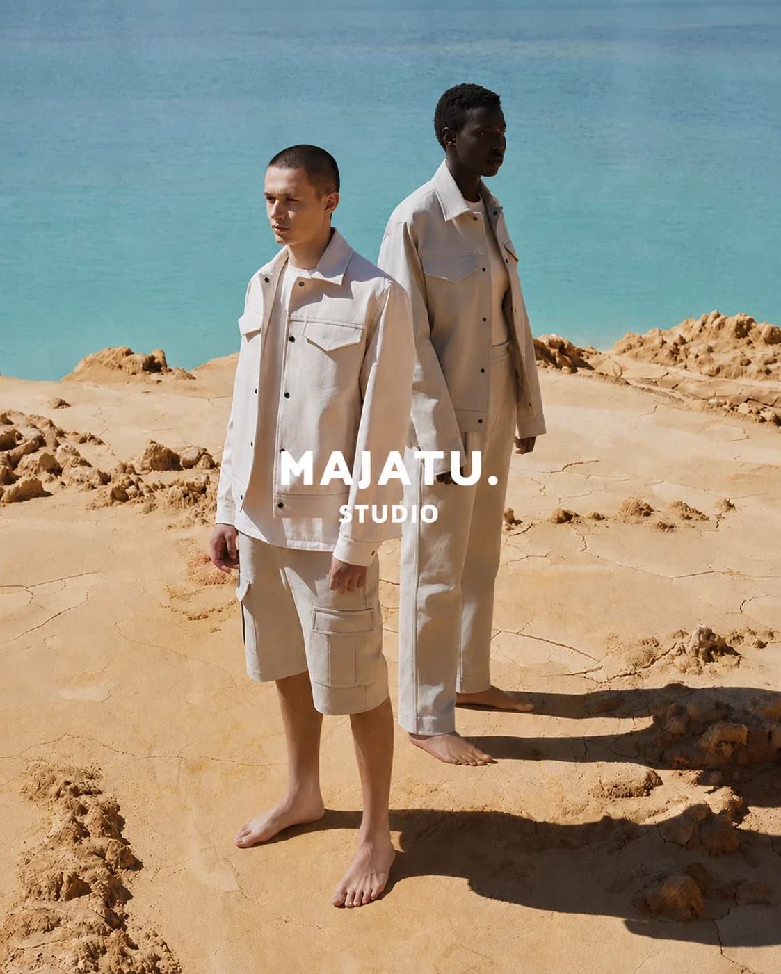 Two models in cream shirting on ochre rock above the sea